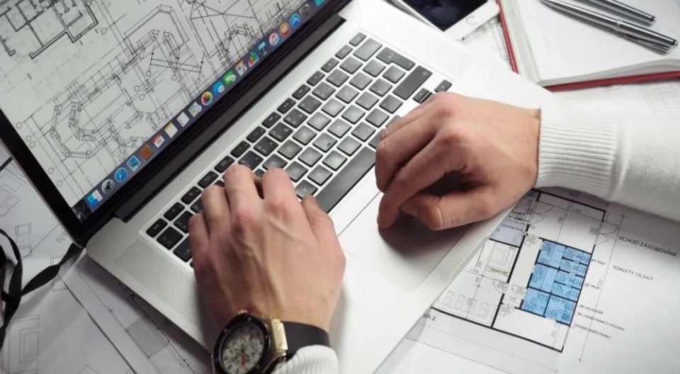 Mani che lavorano su un portatile appoggiato a dei disegni tecnici: progettare le pagine di un sito è come progettare una pianta